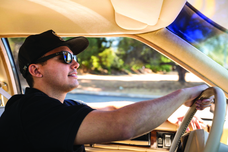 A Millenial in sunglasses and a baseball cap driving a collector car.