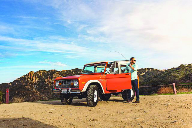 A Millennial parks his classic car on rocky terrain and stands by the door of the driver's side.