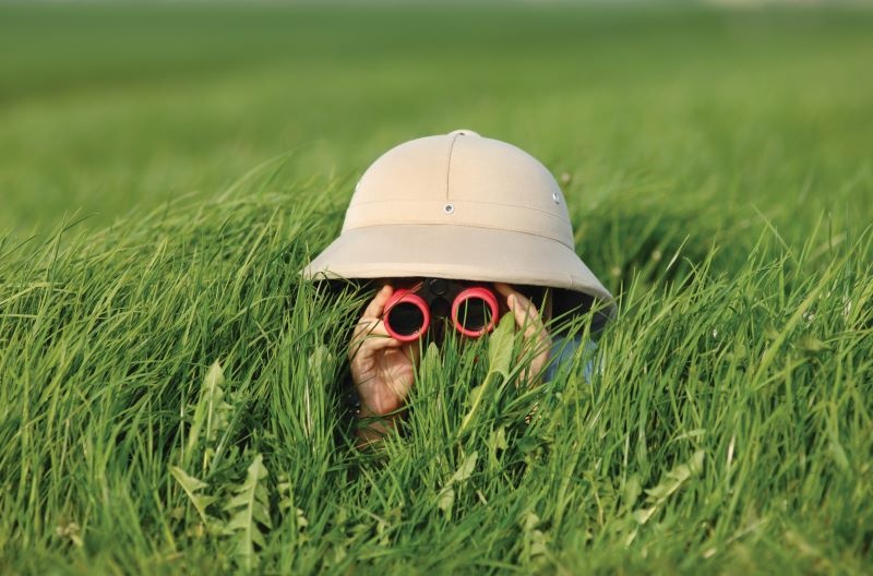 person hiding behind tall grass in bucket hat with binoculars
