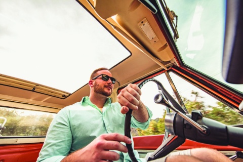 Millennial with sunglasses on driving a collector car with his hands on the steering wheel.
